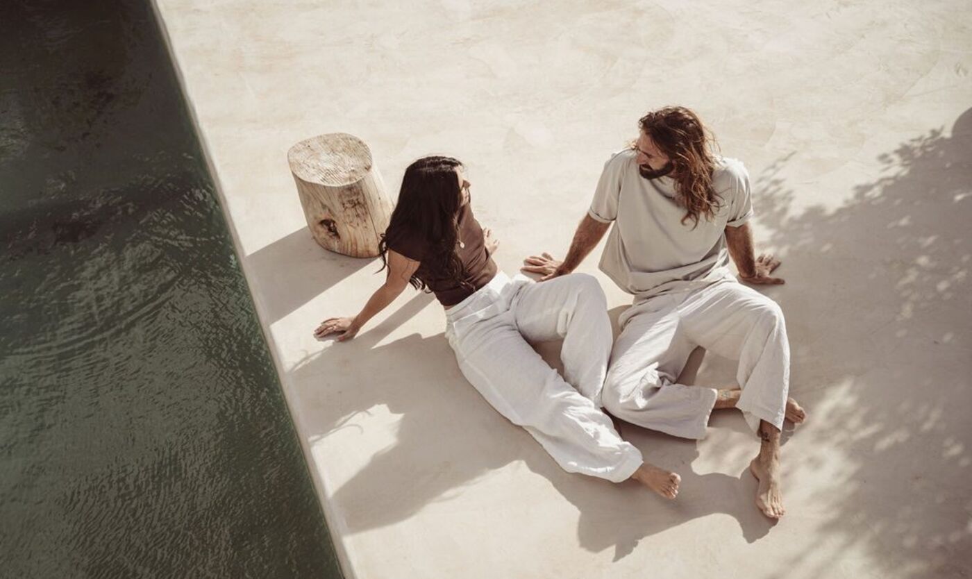 Two people sitting on a sunlit terrace floor in conversation, seen from above, the kind of unhurried talk a clear request needs