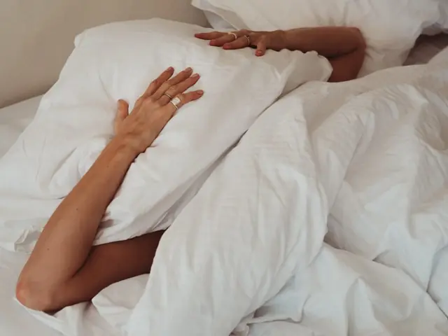 Eine Person in einem Bett mit weißer Bettwäsche, der Kopf unter Kissen und Decke verborgen, eine Hand ruht auf dem Kissen, die andere auf der Decke; nur Arme und Hände sind zu sehen