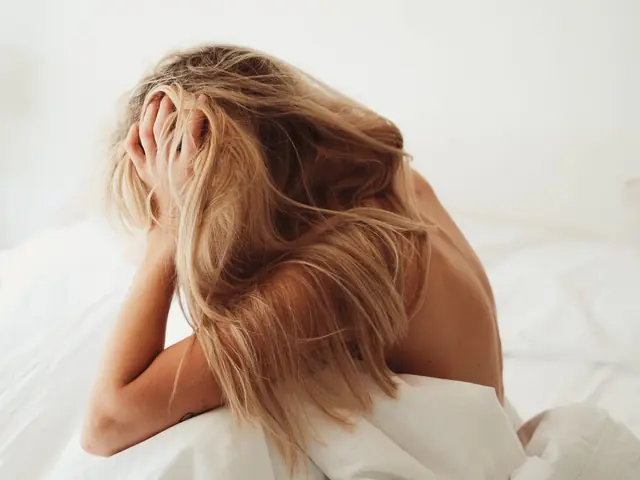 Eine Frau mit zerzaustem blondem Haar sitzt auf einem weißen Bett, den Kopf gesenkt und eine Hand im Haar, mit dem Rücken zur Kamera